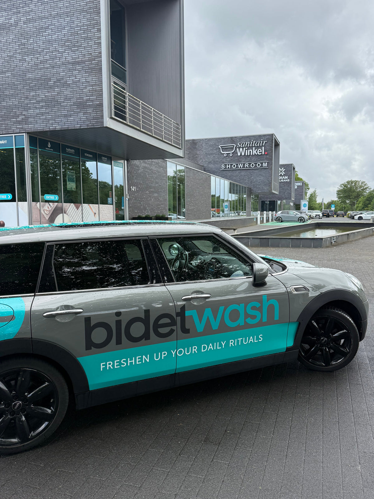 A Bidetwash automatic toilet cleaning vehicle parked on a paved street outside a modern building with the Bidetwash logo visible on the side.